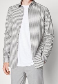 Camisa de botones a rayas en gris y blanco, con mangas largas, un collar y un ajuste slim. Llevada sobre una camiseta blanca lisa.