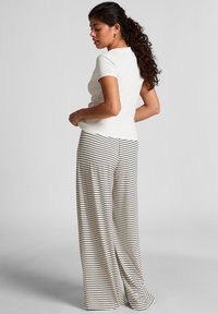 Vrouw met krullend haar in een witte top met korte mouwen en brede zwart-wit gestreepte broek, staand en naar de zijkant kijkend.