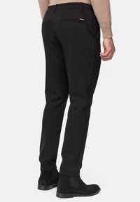 Mężczyzna ubrany w czarne spodnie o prostym kroju, czarne zamszowe botki Chelsea i beżowy sweter, pokazany od tyłu na jednolitym tle.