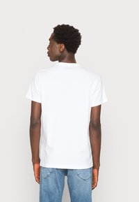 Camiseta de algodón blanca con cuello redondo y mangas cortas, mostrada desde la parte de atrás. Un par de jeans de mezclilla azul claro son visibles a la altura de la cintura.