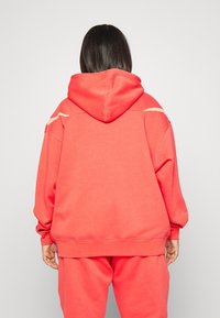 Koralröd oversized hoodie tillverkad av mjuk bomullsblandning, med huva och ribbade muddar, med diskreta elfenbensvita detaljer på axlarna.