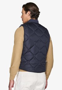 Marineblauer gesteppter Weste mit hohem Kragen, ausgestattet mit einem Rautenmuster und einer einheitlichen Farbe, kombiniert mit einem hellbraunen strukturierten Pullover darunter.