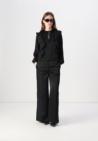 Blusa nera con dettagli arricciati e chiusura a legatura, abbinata a pantaloni neri larghi. Tessuto liscio con una leggera lucentezza, indossata con occhiali da sole neri.