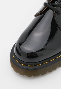 Černý lakovaný kožený shoe s kulatou špičkou, žlutým prošíváním a silnou, strukturovanou podrážkou. Obsahuje černé tkaničky a hladký povrch.