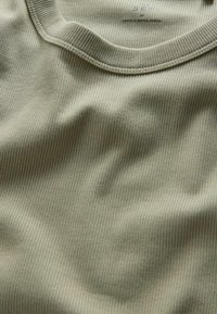 Detailný záber na svetlo olivovú ribbed tkaninu s okrúhlym výstrihom a potlačenou etiketou s nápisom "Vyrobené v Bangladéši" a veľkosťou "S/M".