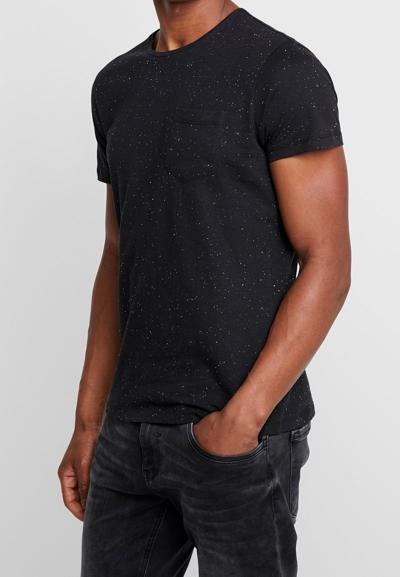 Camiseta negra de manga corta con un patrón salpicado, escote redondo y un bolsillo en el pecho. Fabricada en suave tejido de algodón. Combinada con jeans oscuros.
