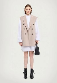 Μπεζ διπλόκουμπο γιλέκο με μεγάλα μαύρα κουμπιά, φορεμένο πάνω από ένα λευκό φόρεμα. Μαύρες μπότα μέχρι τον αστράγαλο και μια μικρή μαύρη τσάντα ολοκληρώνουν την εμφάνιση.