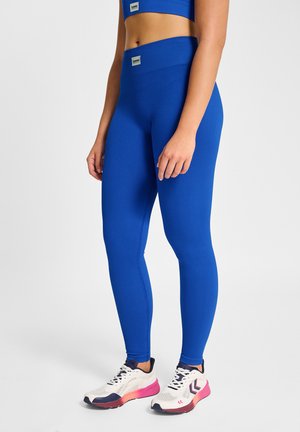 Kobieta nosząca jaskrawo niebieskie prążkowane legginsy, białe buty sportowe z różowymi podeszwami, stojąca na tle jednolitego jasnego tła.