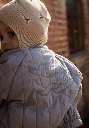 Niño pequeño con un gorro de punto beige con diseños de pájaros y una chaqueta acolchada azul claro con una ballena cosida en la espalda, de espaldas al aire libre.