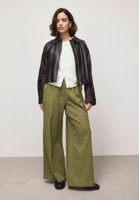 Veste courte en cuir noir portée sur un pull blanc texturé à boutons, associée à un pantalon taille haute à jambes larges à carreaux verts et des chaussures noires.