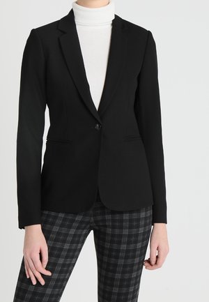 Kvinde iført en tætsiddende sort blazer over en hvid rullekrave, kombineret med sort-grå ternede bukser, stående mod en ensfarvet baggrund.