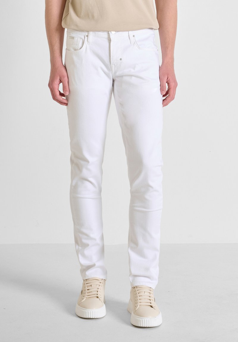 Jeans in denim bianco con vestibilità slim, dotati di cinque tasche e una texture liscia. Abbinati a sneaker beige chiaro.