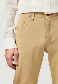 Beige denimbroek met een rechte snit, voorzien van zijzakken, subtiele stiksels en een sluiting met een bovenste knoop. Gecombineerd met een witte linnen blouse.