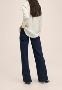 Blouse crème avec manches retroussées et jean large marine, présentant une texture lisse et des poches arrière. Port é avec des chaussures à talons noires.