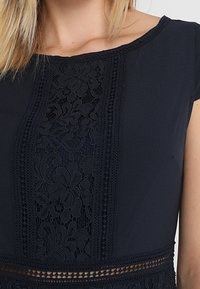 Námořnická modrá šaty s krajkovými detaily na přední straně, s složitými květinovými vzory a texturovanými akcenty podél výstřihu a lemování.