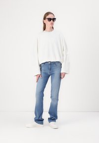 Fehér, oversized, hosszú ujjú ing, világoskék farmer és fehér sportcipő. A modellen fekete napszemüveg van; minimális háttér.