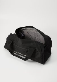 Black duffel bag made from durable material, featuring a zip closure, two sturdy handles, and a simple logo on the side. Interior is gray.