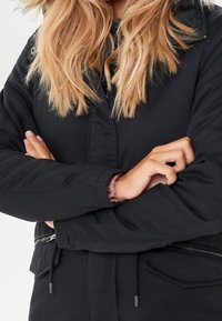 Giacca nera con tasca con zip, cordini in vita e un cappuccio morbido e testurizzato. Tessuto liscio con vestibilità comoda.