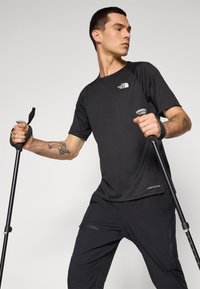 Camisola atlética preta de manga curta com decote redondo e logótipo, vestida com calças pretas, segurando bastões de trekking, com um tecido leve.