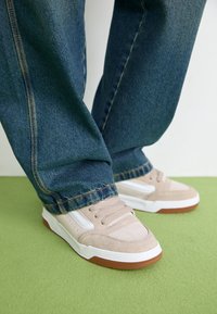 Beige semskede joggesko med hvite og brune detaljer, kombinert med lyseblå jeans, på en grønn overflate. Joggeskoene har rund tå og snøring.