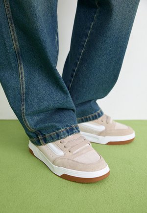 Beige semskede joggesko med hvite og brune detaljer, kombinert med lyseblå jeans, på en grønn overflate. Joggeskoene har rund tå og snøring.