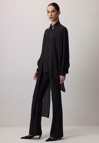 Czarna koszula z długim rękawem o luźnym kroju, z zapięciem na guziki i ozdobnym kołnierzykiem. W połączeniu z dopasowanymi czarnymi spodniami.