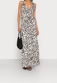 Robe maxi à base crème avec un motif zèbre noir, présentant un col en V, sans manches et une taille ceinturée. Sac à main noir et sandales.