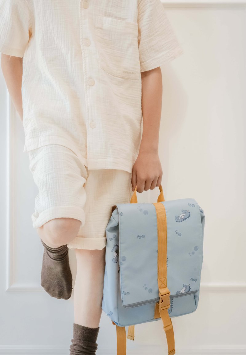 Modra nahrbtnik z vzorcem morskega konjička, oranžnimi trakovi in sponko. Otrok nosi svetlo srajco z gumbi in bež kratke hlače ter drži torbo.
