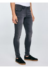 Férfi, szűk, kifakult fekete farmerben és gumitalpú fekete sportcipőben, világos, egyszínű háttér előtt áll.