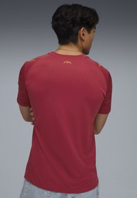 Röd kortärmad T-shirt med en slät textur. Har guld-vampyrlogotyper på axelpartierna. Avslappnad design, rundad halsringning och ledig passform.