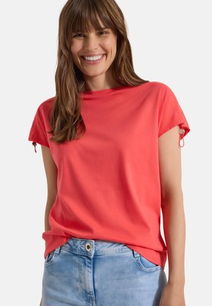 Smilende kvinde med langt brunt hår bærer en lys koral kortærmet top og lyseblå jeans mod en ensfarvet baggrund.
