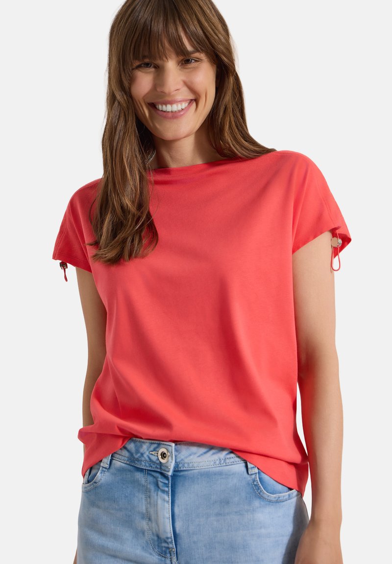 Femme souriante aux longs cheveux bruns portant un haut corail vif à manches courtes et un jean bleu clair sur un fond uni.