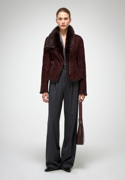 Mujer de pie con una chaqueta de cuello de piel marrón oscuro, pantalones anchos grises, zapatos negros y un bolso marrón a juego en la mano.