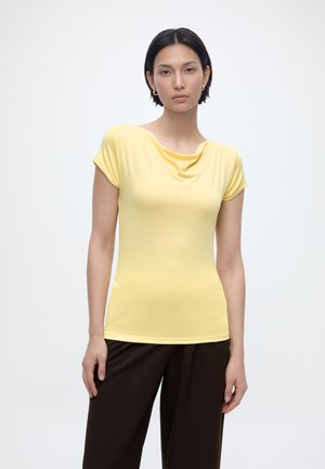 Jeune femme debout, portant un haut à manches courtes jaune clair et un pantalon marron foncé, avec des cheveux courts noirs et des boucles d'oreilles en or en forme de créoles.