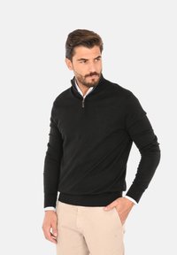 Maglione nero in maglia con collo a zip a un quarto, polsini e orlo a coste. Il modello lo indossa sopra una camicia bianca con colletto, abbinato a pantaloni beige.