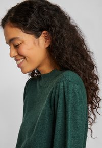 Groene trui met hoge hals, textuurstof en geplooide schouderdetails. Zacht, comfortabel uiterlijk met een relaxte pasvorm.