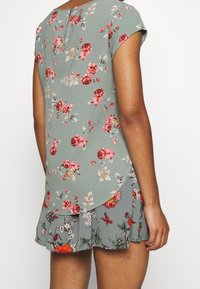 Top floreale in verde polveroso con maniche corte, orlo curvo e dettaglio a chiave sul retro. Il tessuto presenta motivi floreali in rosa e rosso.