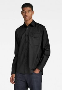 Camisa negra de manga larga abotonada con un bolsillo en el pecho, hecha de algodón suave. Cuenta con un cuello clásico y un sutil detalle de la marca en el bolsillo.