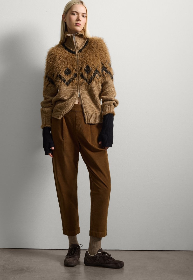 Felpa zip-up marrone con colletto peloso e motivo geometrico nero, abbinata a pantaloni di velluto a coste beige e scarpe scure. Dettagli del tessuto testurizzati visibili.