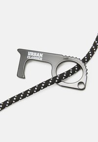 Svart metallverktyg med handtag och tandad kant, försett med texten "URBAN CLASSICS," tillsammans med ett svart och vitt flätat rep.