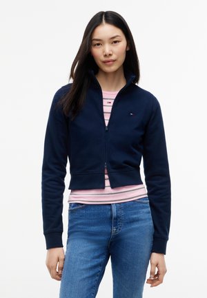 Ung kvinde med langt mørkt hår iført en navy lynlåsjakke over en lyserød stribet skjorte og blå jeans mod en ensfarvet hvid baggrund.