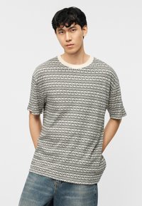 Νέος άνδρας που φοράει πουλόβερ με μοτίβο και κοντά μανίκια και μπλε τζιν με τα χέρια στις τσέπες, μπροστά σε απλό ανοιχτό φόντο.