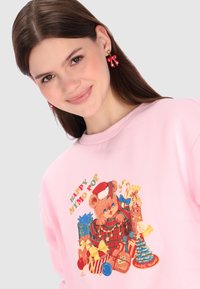 Sudadera rosa con un gráfico de oso con regalos y elementos navideños. Pendientes de lazo rojo que añaden un toque festivo. Textura de tela suave.