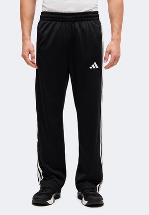 Άνδρας που φοράει μαύρο παντελόνι Adidas με λευκές ρίγες στα πλάγια και μαύρα αθλητικά παπούτσια, στέκεται σε απλό φόντο.