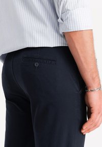 Pantaloni di tessuto blu navy con una superficie liscia e una tasca posteriore singola con chiusura a bottone, abbinati a una camicia a righe blu chiaro.