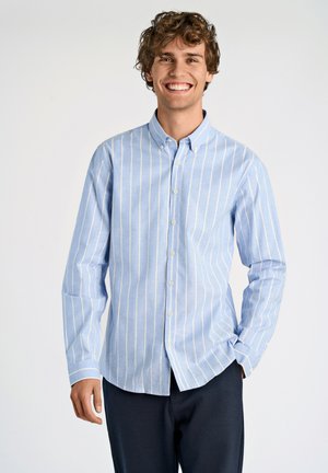 Smilende ung mand med krøllet hår iført en lyseblå, vertikalt striped skjorte og mørke bukser, stående mod en ensfarvet baggrund.