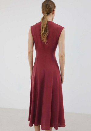 Vrouw met lichtbruin haar in een paardenstaart, die een mouwloze, aansluitende donkerrode midi-jurk draagt, met de rug naar een effen lichte achtergrond gekeerd.