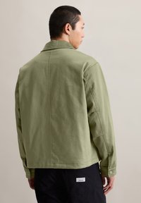 Hellgrüne Baumwolljacke mit strukturierter Silhouette, Rückennaht-Detail und geknöpften Manschetten, kombiniert mit schwarzen Hosen mit einem Etikett.