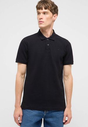 Junger Mann, der ein unifarbenes schwarzes kurzärmeliges Poloshirt und blaue Jeans trägt und nach links blickt vor einfachem Hintergrund.