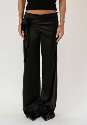 Mujer con pantalones anchos de satén negro de talle alto, con un detalle de nudo atado en la cadera izquierda y zapatos negros de punta.